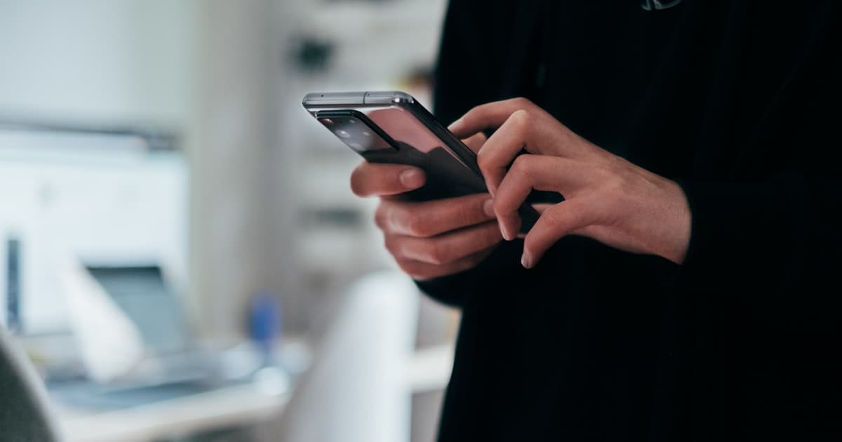 A person typing a message on their smartphone with a laptop open in the background